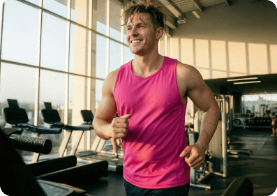 A smiling white man in a pink tank top running on a treadmill in a sunlit gym