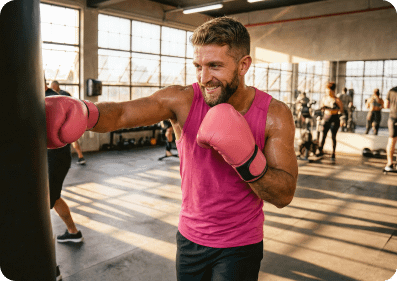 A smiling white man with pink boxing gloves punches a heavy bag in a sunlit brick gym.
