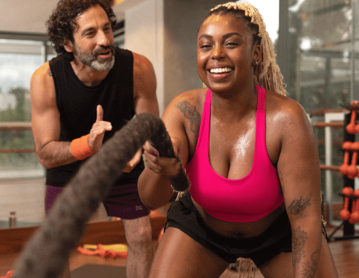 A woman is exercising using ropes, with her personal trainer encouraging from behind.