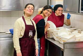 Three smiling wellhubbers volunteers in maroon aprons stack bowls in a professional kitchen during a Global Volunteer Day event.