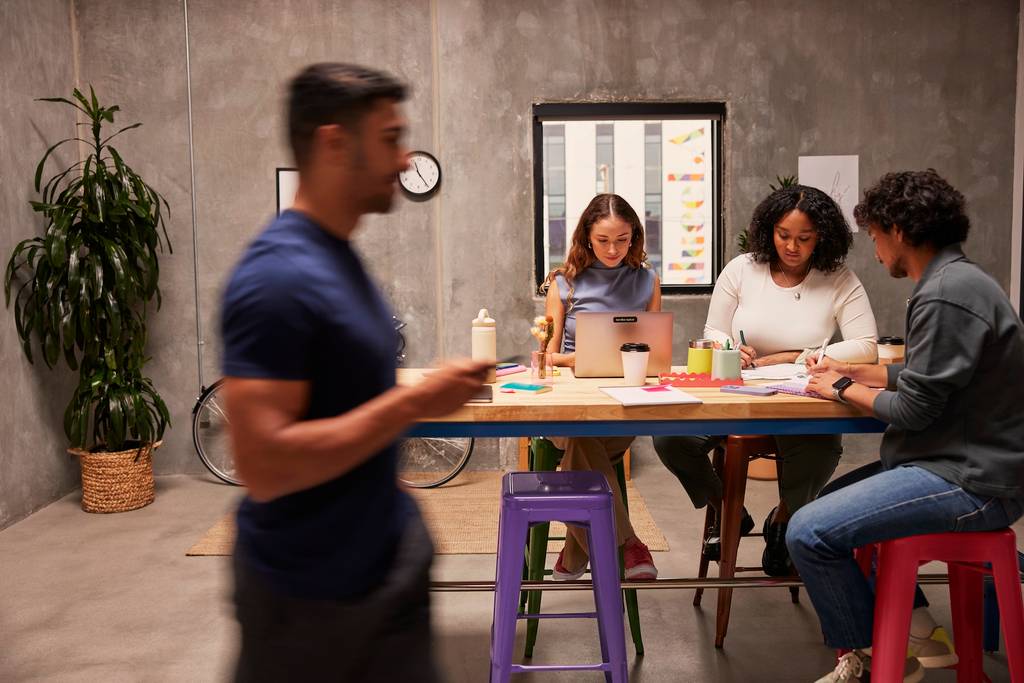 Four people at an office, sitting down and standing up, with office supplies and computers.