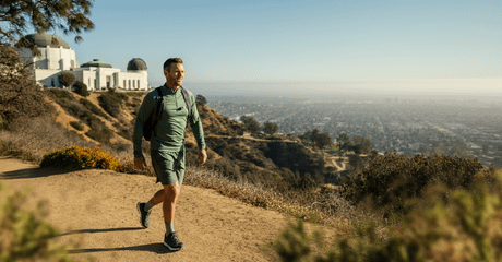 Homem caminha por uma trilha de terra em uma colina, usando mochila e roupas esportivas, com vista panorâmica da cidade ao fundo e um edifício com cúpulas ao lado esquerdo.