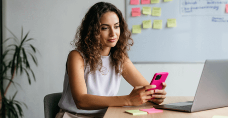 Mulher sentada à mesa olhando o celular rosa, com um laptop aberto à frente e um quadro branco ao fundo com post-its coloridos.