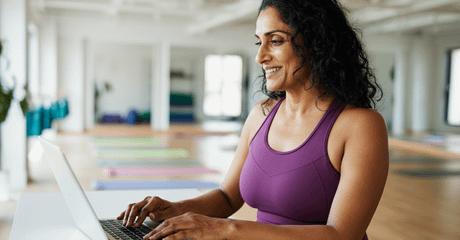 Mulher usando notebook em estúdio de yoga amplo e iluminado, com tapetes alinhados ao fundo. Ela veste roupa esportiva roxa e está sentada à mesa digitando no computador.