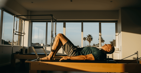 Homem deitado em um aparelho de Pilates, com joelhos dobrados e pés apoiados, realizando exercício em uma sala iluminada por grandes janelas com vista para palmeiras.