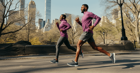 Duas pessoas correndo lado a lado em uma via asfaltada de parque urbano, vestindo roupas esportivas roxas, com árvores sem folhas e prédios altos ao fundo.