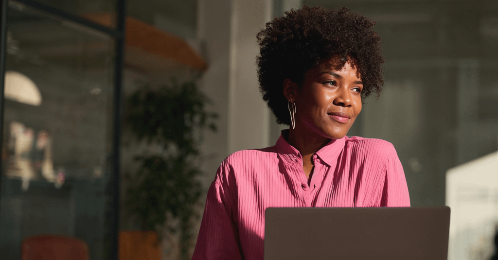 Mulher vestindo uma camisa rosa sentada com um laptop em um ambiente de escritório. Ela está iluminada por luz natural, olhando de forma pensativa e sorridente para o lado.
