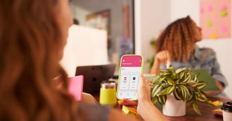 Mulher segurando um smartphone com um app aberto durante uma reunião informal; outra mulher ao fundo com laptop e post-its coloridos na parede.