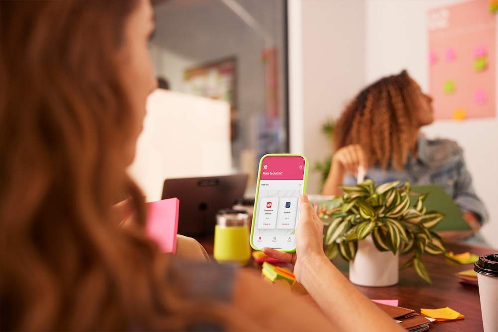 Mulher segurando um smartphone com um app aberto durante uma reunião informal; outra mulher ao fundo com laptop e post-its coloridos na parede.