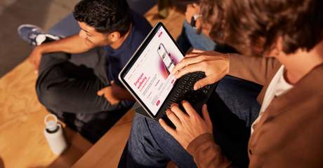 Pessoa usando um tablet com teclado no colo, navegando por um site sobre bem-estar no trabalho, sentada em degraus de madeira com outras pessoas ao redor.