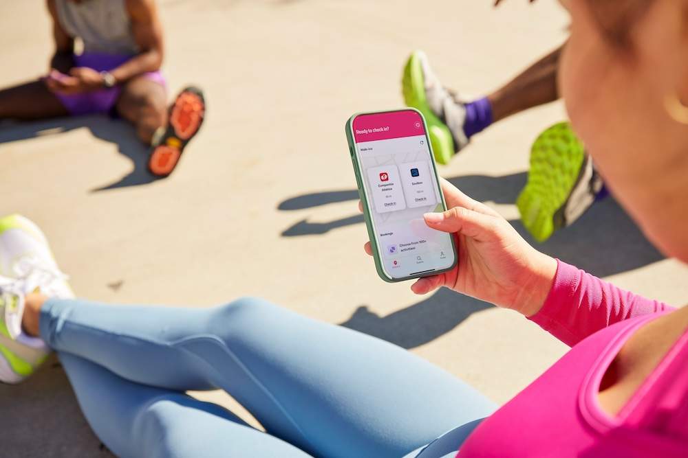Pessoa em roupa de ginástica segura um smartphone com um app de check-in de treino, sentada no chão ao ar livre, com outras pessoas em trajes esportivos ao fundo.
