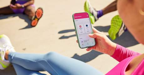 Pessoa em roupa de ginástica segura um smartphone com um app de check-in de treino, sentada no chão ao ar livre, com outras pessoas em trajes esportivos ao fundo.