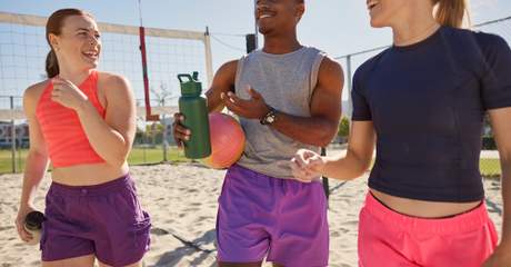 Três pessoas sorridentes conversando em uma quadra de vôlei de praia, segurando garrafas e bola, representando socialização, atividade física e bem-estar.