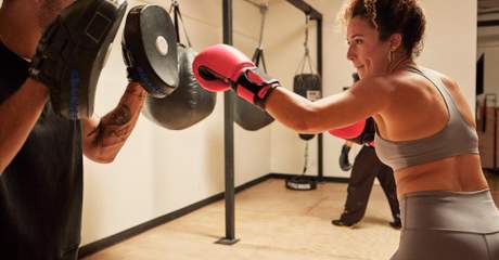 Pessoa usando luvas de boxe rosas treina golpes com um instrutor que segura manoplas em uma academia, com sacos de pancada pendurados ao fundo.