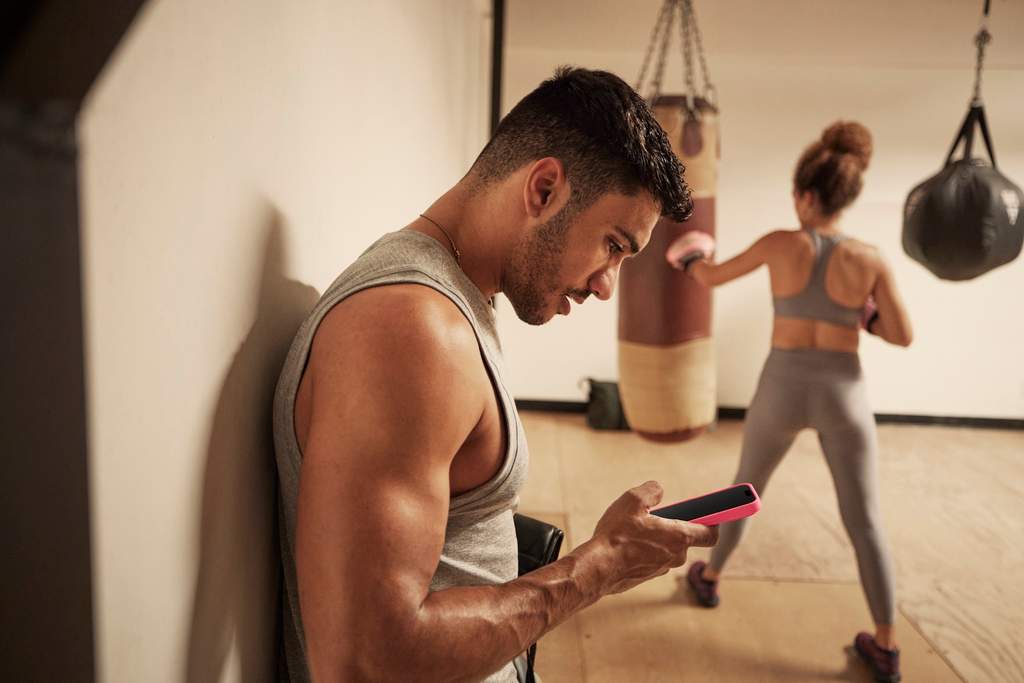 Homem encostado na parede consultando o celular em uma academia, enquanto ao fundo uma mulher treina golpes em um saco de boxe.