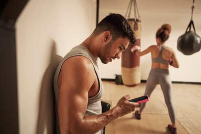 Homem encostado na parede consultando o celular em uma academia, enquanto ao fundo uma mulher treina golpes em um saco de boxe.