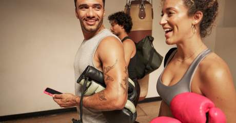 Homem e mulher sorrindo após aula de boxe, segurando luvas e conversando de forma descontraída em uma academia, com saco de pancadas ao fundo.