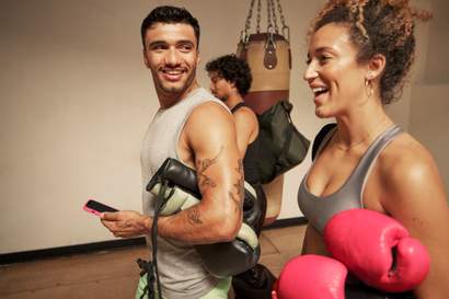 Homem e mulher sorrindo após aula de boxe, segurando luvas e conversando de forma descontraída em uma academia, com saco de pancadas ao fundo.