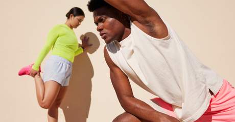 Homem e mulher fazem alongamento ao ar livre em frente a uma parede clara; ele se inclina para o lado com o braço estendido, enquanto ela alonga a perna apoiada na mão.