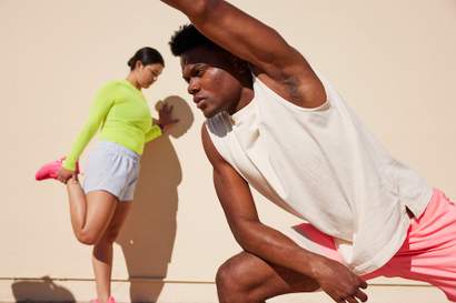 Homem e mulher fazem alongamento ao ar livre em frente a uma parede clara; ele se inclina para o lado com o braço estendido, enquanto ela alonga a perna apoiada na mão.