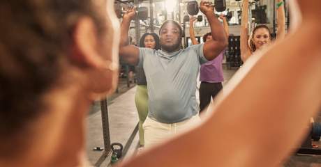 Grupo de pessoas em uma academia, levantando halteres acima da cabeça durante treino funcional em circuito.