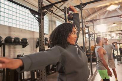 Mulher levantando um kettlebell com uma mão em uma academia, com outras pessoas se exercitando ao fundo.