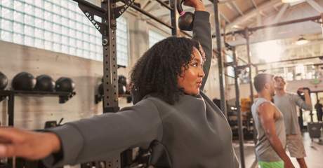 Mulher levantando um kettlebell com uma mão em uma academia, com outras pessoas se exercitando ao fundo.