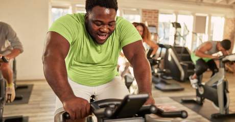 Homem sorridente se exercitando em bicicleta ergométrica durante aula de spinning em academia, com outras pessoas ao fundo.