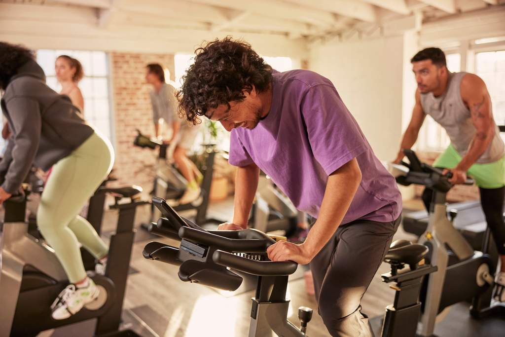 Pessoas participam de uma aula de ciclismo indoor em uma academia iluminada, pedalando em bicicletas estacionárias lado a lado.