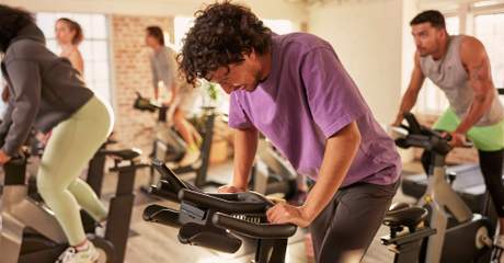 Pessoas participam de uma aula de ciclismo indoor em uma academia iluminada, pedalando em bicicletas estacionárias lado a lado.