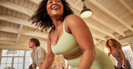 Mulher sorridente praticando ciclismo indoor em grupo, em um ambiente iluminado e descontraído, promovendo saúde, bem-estar e atividade física.