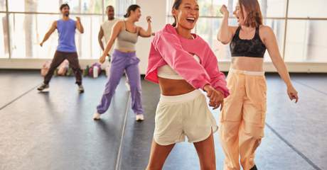 Grupo de pessoas sorrindo e se divertindo durante uma aula de dança em estúdio, movimentando-se de forma enérgica e descontraída.