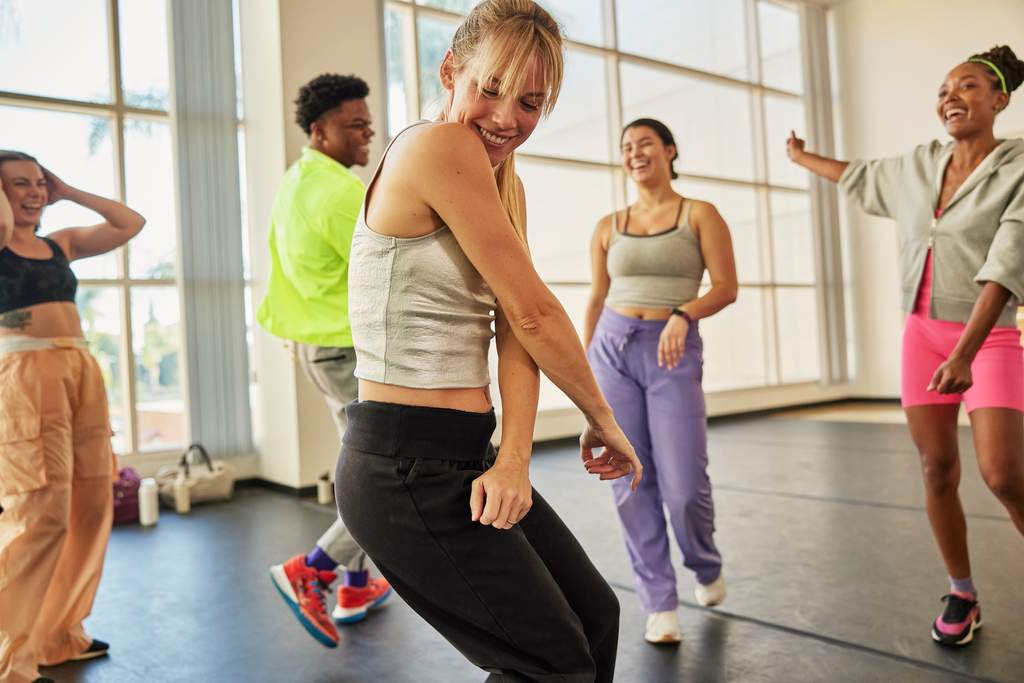 Grupo de pessoas se movimentando em uma aula de dança em estúdio iluminado, vestindo roupas esportivas e aparentando estar engajadas na atividade.