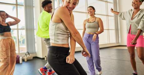 Grupo de pessoas se movimentando em uma aula de dança em estúdio iluminado, vestindo roupas esportivas e aparentando estar engajadas na atividade.