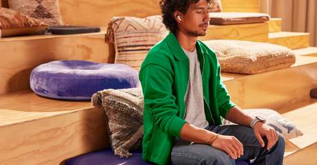 Pessoa sentada em um banco acolchoado com almofadas ao redor, usando fones de ouvido e olhos fechados, em um ambiente de madeira claro e acolhedor.