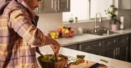 Homem preparando salada fresca em casa, misturando vegetais em tigela de madeira sobre bancada de cozinha.