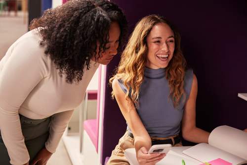 Duas mulheres colaboram alegremente em uma mesa; uma está sentada com celular e caderno aberto, enquanto a outra se inclina para participar.