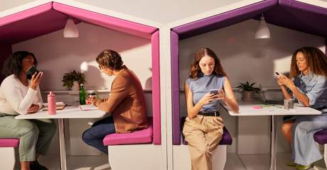 Quatro pessoas em um ambiente de trabalho moderno, sentadas em cabines acolchoadas, conversando e usando celulares e notebooks, representando diferentes formas de comunicação no escritório.