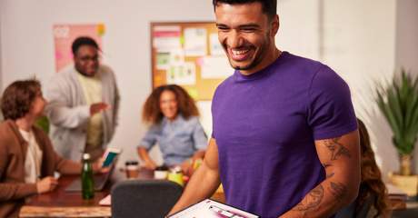Homem sorridente segurando um tablet em uma sala de reunião, com colegas conversando ao fundo em um ambiente de trabalho colaborativo.