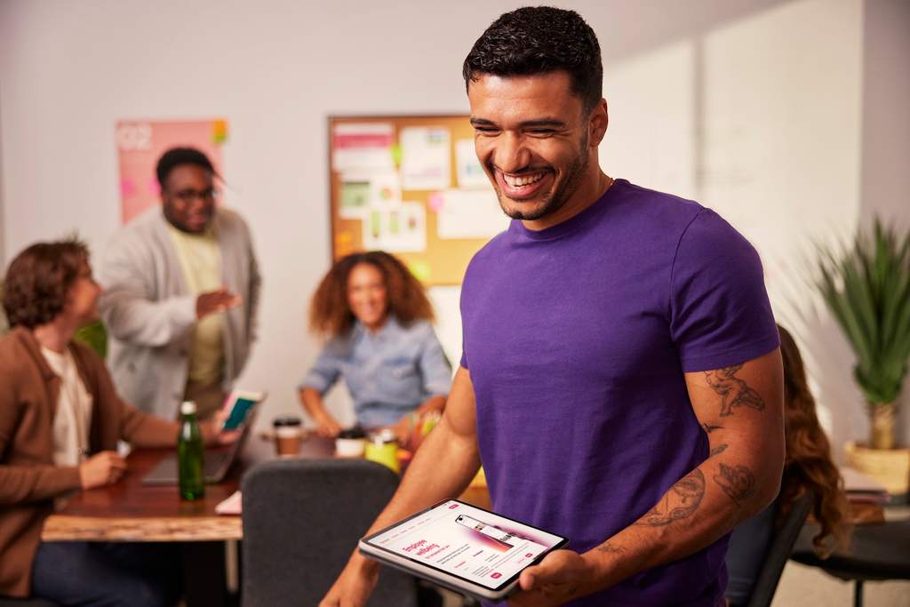 Homem sorridente segurando um tablet em uma sala de reunião, com colegas conversando ao fundo em um ambiente de trabalho colaborativo.
