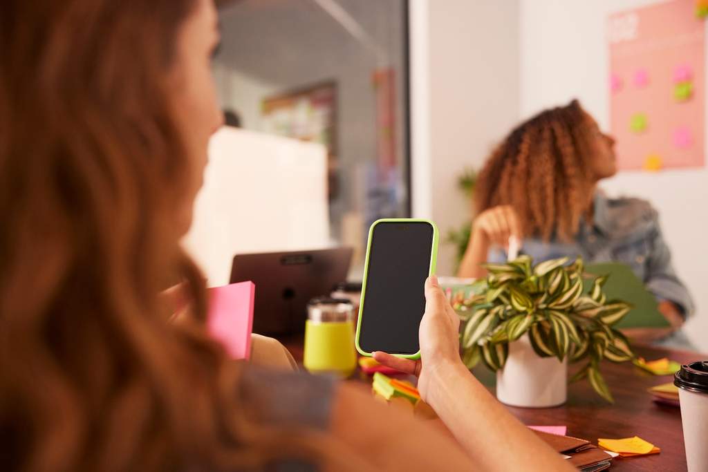 Pessoa segurando um smartphone em uma reunião de trabalho, com colegas ao fundo e mesa com laptop, post-its e planta decorativa.