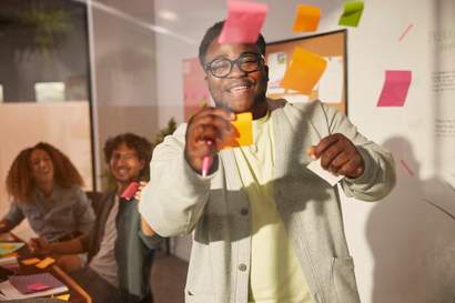 Homem sorrindo enquanto coloca notas adesivas coloridas em uma parede de vidro durante uma reunião, com dois colegas rindo ao fundo.