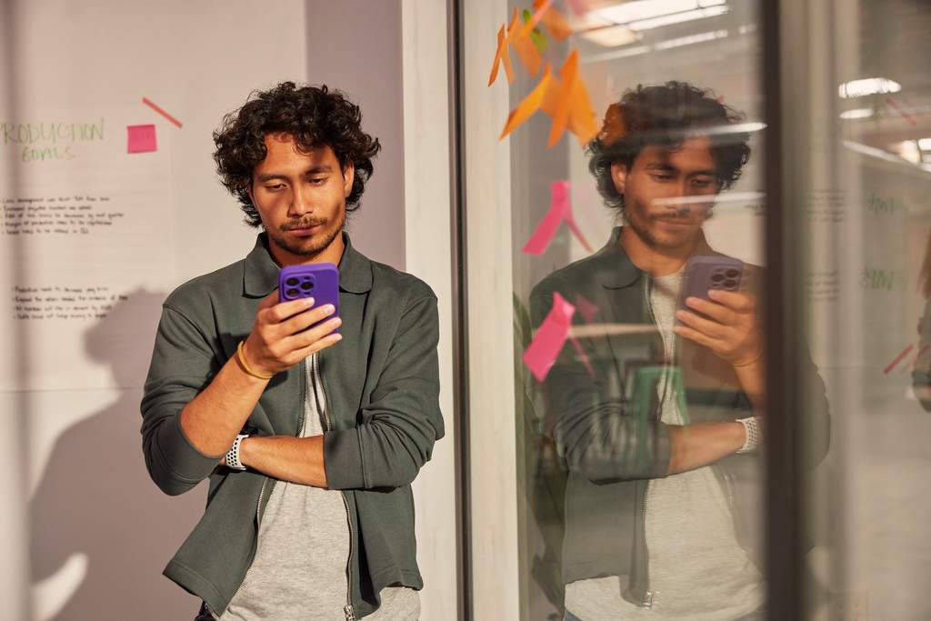 Pessoa em pé, de frente para um vidro com post-its coloridos, segura um celular roxo e olha para a tela com atenção.