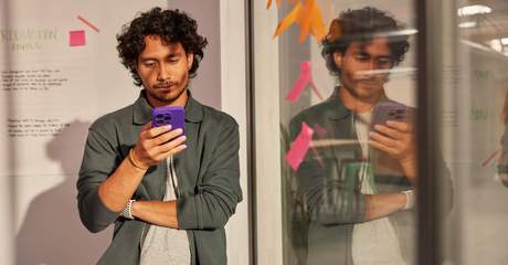 Pessoa em pé, de frente para um vidro com post-its coloridos, segura um celular roxo e olha para a tela com atenção.