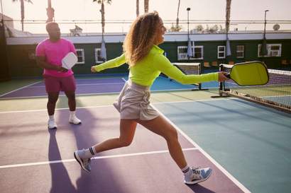 Duas pessoas jogando pickleball em uma quadra ao ar livre; a jogadora em primeiro plano retorna a bola com uma raquete amarela enquanto o jogador ao fundo observa preparado.