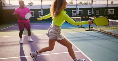 Duas pessoas jogando pickleball em uma quadra ao ar livre; a jogadora em primeiro plano retorna a bola com uma raquete amarela enquanto o jogador ao fundo observa preparado.