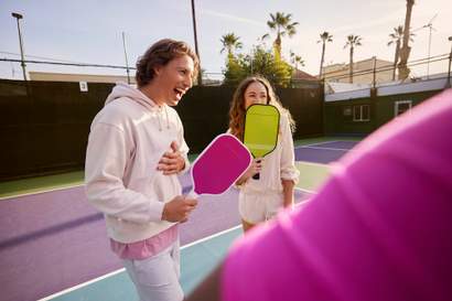 Duas pessoas em uma quadra de pickleball seguram raquetes coloridas e sorriem, com o sol se pondo ao fundo e palmeiras ao redor da quadra.