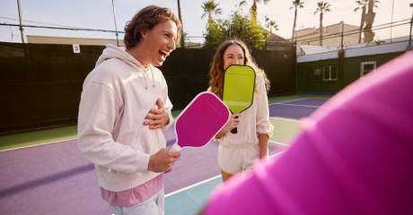 Duas pessoas em uma quadra de pickleball seguram raquetes coloridas e sorriem, com o sol se pondo ao fundo e palmeiras ao redor da quadra.