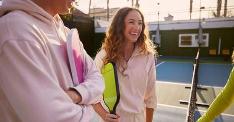 Grupo de jovens sorrindo e conversando em uma quadra de pickleball ao ar livre, segurando raquetes coloridas, em um ambiente ensolarado e descontraído.