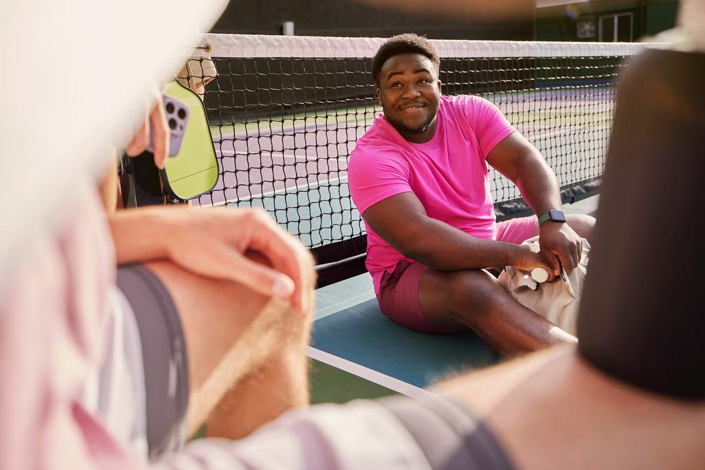Homem sorridente, vestido com camiseta e shorts rosa, sentado em uma quadra de tênis depois de jogar, representando bem-estar, socialização e prática esportiva.
